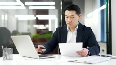 Meşgul Asyalı işadamı, iş yerinde oturan bir dizüstü bilgisayarla evrak işleriyle uğraşıyor. Resmi takım elbiseli, kendine güvenen olgun bir adam evrakları dolduruyor, yazıyor, iş yerinde bankacılık yapıyor.