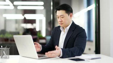 Asyalı hayal kırıklığına uğramış bir adamın iş yerinde oturan dizüstü bilgisayarında yazılımla veya servisle ilgili sorunları var. Resmi takım elbiseli endişeli bir işadamı kötü internet bağlantısından şikayet ediyor