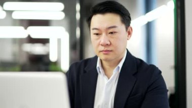 Ciddi Asyalı işadamı iş yerinde dizüstü bilgisayar kullanıyor. Resmi bir takım elbise sohbetine odaklanmış bir adam online, bankacılık, bilgisayar uygulamasında çalışıyor ya da bir başlangıç projesi düşünüyor. Kapat.