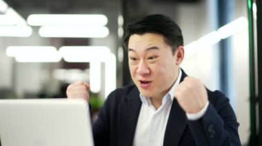 Mutlu Asyalı işadamı iş yerindeki bilgisayarından harika haberler aldı. Resmi takım elbiseli, şaşırmış gülümseyen adam bilgisayardan olumlu bir mesaj okuyor. Kapat.