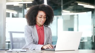 İş yerindeki bir laptopta oturan takım elbiseli Afro-Amerikan bir iş kadınının portresi. Gözlüklü, gülümseyen siyah kadın kameraya bakıyor. Kadın yöneticinin vesikalığı.