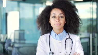 Modern hastane kliniğinde kameraya bakan beyaz önlüklü genç, mutlu Afro-Amerikan kadın doktor portresi. Gülümseyen siyah kadın doktor ofiste poz veriyor. Kapat.