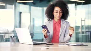 Mutlu Afro-Amerikalı iş kadını internet üzerinden alışveriş yapıyor. İş yerinde telefonunda kredi kartı numarasını yazıyor. Gülümseyen siyah kadın indirimlerden ve terfilerden zevk alıyor.