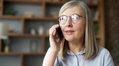 Yaşlı gri saçlı kadın evdeki oturma odasında kanepede otururken telefonla konuşuyor. Yaşlı bir kadın iş arkadaşıyla konuşuyor ya da akıllı telefon kullanarak ailesiyle iletişim kuruyor. Kapat.