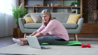 Son sınıf bayan, evde oturma odasındaki paspasın üzerinde oturmuş dizüstü bilgisayarla jimnastik yapıyor. Yaşlı bir kadın video aracılığıyla eğitmeni dinleyerek esneme egzersizleri yapıyor.