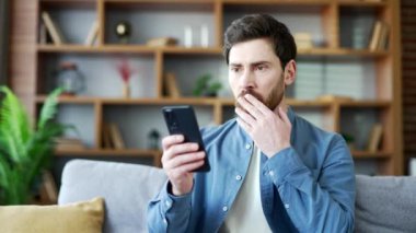 Stresli adam evdeki oturma odasında koltukta otururken cep telefonundan kötü haberler okuyor. Üzgün sakallı erkek üzgün ve üzüntüden ağlıyor. Akıllı telefondaki olumsuz mesajı gözden geçiriyor.