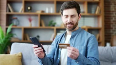 Mutlu, sakallı, online alışveriş yapan, cep telefonunda kredi kartı numarası yazan, evdeki kanepede oturan yakışıklı adam. Gülümseyen erkek indirimler ve terfilerle tatmin olan bir alışveriş yapar. Kapat.