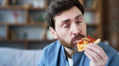 Oturma odasındaki kanepede oturmuş pizza yiyen neşeli yakışıklı adam. Gülümseyen sakallı olgun bir erkek hızlı yemek teslimatı konusunda mutlu. Eve yemek götürmek için sipariş veriyor. Kapat.