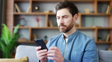 Evdeki oturma odasında koltukta oturan cep telefonu kullanan yakışıklı adam. Gülümseyen sakallı bir erkek sosyal ağlara takılır, online alışveriş yapar, sohbet eder, akıllı telefondan mesajlar yazar veya okur. Kapat. 