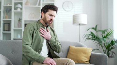 Göğüs tansiyonundan muzdarip hasta bir adam, evdeki oturma odasında kanepede oturan kalp krizi. Olgun sakallı erkek elleriyle göğsünü tutuyor, derin nefes alıyor, acil tıbbi müdahaleye ihtiyacı var.