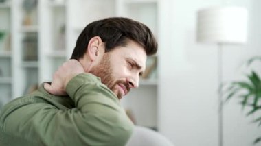Evdeki kanepede otururken boynu ağrıyan yorgun bir adam. Hasta, olgun, sakallı erkek masajı elleri ve kasları ağrıyor, esniyor, boynunun arkasını ovuyor. Kapat.