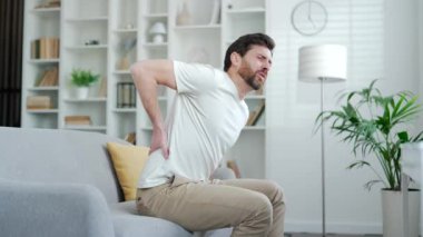 Evdeki oturma odasında kanepede otururken sırt ağrısı çeken yorgun bir adam. Üzgün olgun sakallı erkek, elleriyle belini tutuyor, masaj yapıyor, kasları ağrıyor, siyatiği ya da hasta omurgası var.