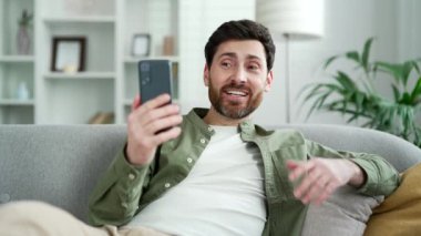 Video görüşmesinde cep telefonu kullanarak konuşan mutlu yakışıklı adam evdeki oturma odasındaki kanepede uzanmış selfie çekiyor. Gülümseyen pozitif sakallı erkek bir arkadaşıyla ya da meslektaşıyla internette sohbet ediyor.