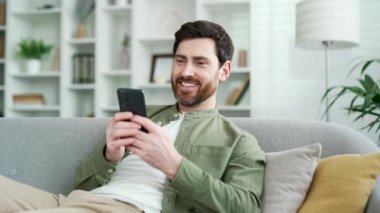 Evdeki oturma odasındaki rahat koltukta cep telefonu kullanan mutlu yakışıklı adam. Gülümseyen erkek sosyal ağlara takılır, internette sohbet eder, ilginç bir şey izler, akıllı telefondan mesajlar yazar.
