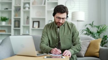 Kulaklık takmış bir iş adamı online video konferansı izliyor. Not alıyor. Ev ofisinde oturan dizüstü bilgisayara bakıyor. Uzaktan kumandalı iş eğitimi alan ya da özel öğretmenle konuşan bir adam.