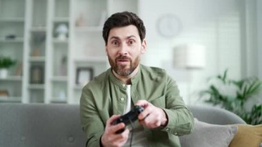 Evdeki oturma odasında kanepede otururken oyun oynayan mutlu ve heyecanlı bir adamın ön görüntüsü. Neşeli yakışıklı erkek ilginç oyun kumandasını elinde tutmaktan zevk alıyor.