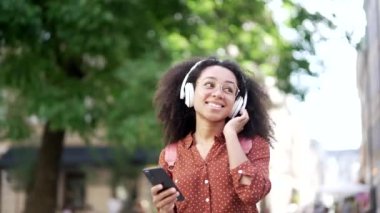 Neşeli, Afro-Amerikalı genç bir kadın. Kablosuz kulaklıkla müzik dinliyor. Şehir caddesinde yürürken akıllı telefon kullanıyor.