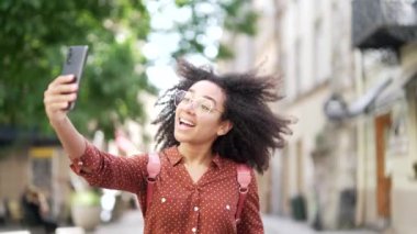Afro-Amerikalı genç bayan turist ya da öğrenci şehir caddesinde yürürken cep telefonuyla arkadaşlarıyla konuşuyor ve el sallıyor. Açık havada akıllı telefon kullanıyor ve internet üzerinden iletişim kuruyor.