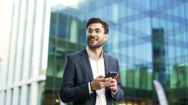 Takım elbiseli kendine güvenen işadamı modern cam ofis binasının yakınında elinde akıllı telefon tutarak yürüyor. Gözlüklü adam, girişimci, yatırımcı, yardımcı ya da yönetici dışarıda cep telefonu kullanıyor.