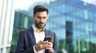 Takım elbiseli kendine güvenen işadamı modern cam ofis binasının yanında elinde akıllı telefonu tutuyor. Gözlüklü adam, girişimci, yönetici ya da yönetici dışarıda cep telefonu kullanıyor.