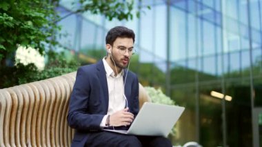 Takım elbiseli bir iş adamı, ofis binasının yanındaki bankta otururken bilgisayarla video görüşmesi yapıyor. Kulaklık takan kendine güvenen girişimci müşteri veya iş ortağıyla çevrimiçi bir toplantı yapıyor.