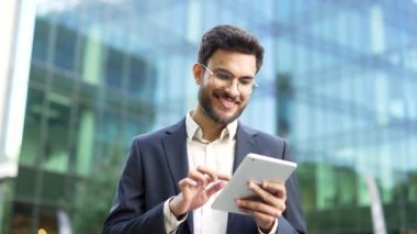 Takım elbiseli kendine güvenen işadamı modern cam ofis binasının yanında masa kullanıyor. Gözlüklü adam, girişimci, yönetici ya da yönetici dışarıda dijital bir cihaz üzerinde çalışıyor.