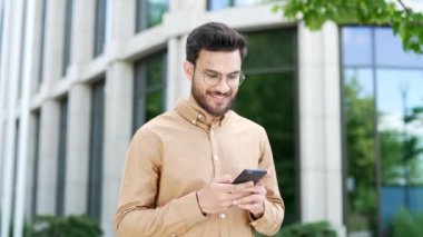 Mutlu iş adamı bir iş yeri binasının yanındaki caddede duran telefonu kullanıyor. Gülümseyen, sakallı, yakışıklı bir erkek internette mesajlaşıyor, internette geziniyor, akıllı telefondan mesajlar yazıyor.
