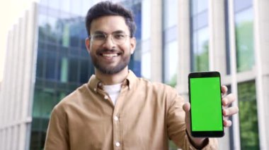 Mutlu yakışıklı iş adamı yeşil ekranlı cep telefonunu bir iş yeri binasının yanında duruyor. Renk anahtarı, reklam için dikey şablon düzeni. Gülümseyen adam kameraya bakıyor.