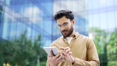 Kendine güvenen işadamı, tablet kullanarak modern cam ofis binasının yanında duruyor. Gözlüklü adam, girişimci, işçi, sahibi ya da yöneticisi dışarıda dijital bir cihaz üzerinde çalışıyor.
