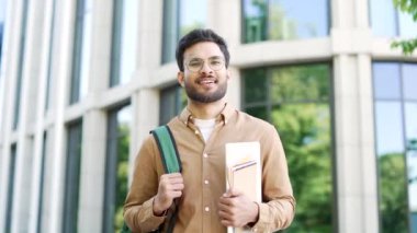 Elinde kitaplarla elinde kitaplarla üniversite binasının yanındaki kampüs boşluğunda kameraya bakan mutlu yakışıklı öğrencinin portresi. Sırt çantalı ve gözlüklü gülümseyen sakallı bir erkek resmi.