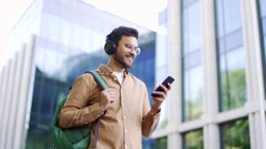 Sırt çantalı yakışıklı bir öğrenci üniversite binasının yanındaki kampüste yürürken akıllı telefon kullanıyor. Arkadaşla online erkek sohbetleri, sosyal ağlara göz atmak, mesajları yazmak veya okumak