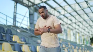 Bir stadyumda sandık tutan genç bir atlet potansiyel kalp krizi belirtileri gösteriyor. Adam güneş ışığı altında sıkıntılı görünüyor, fiziksel aktivite sırasında sağlık riskini vurguluyor..