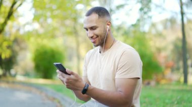 Parkta akıllı telefon kullanarak kulaklık takan gülümseyen genç adam. Müzikten, mesajlaşmadan ve sağlıklı ve aktif yaşam tarzından hoşlanan kişi.