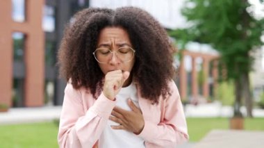 Üniversite binasının yakınındaki caddede öksüren Afrikalı Amerikalı kadın. Soğuk algınlığı ya da grip geçiren genç zenci kadın kendini kötü hissediyor, elini göğsüne dayıyor, zatürreeden ya da virüsten muzdarip. Kapat.