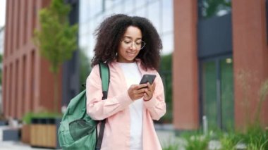 Gülümseyen Afrikalı Amerikalı kız öğrenci üniversite binasının yanındaki kampüs alanında telefon kullanıyor. Sırt çantalı mutlu bir zenci kadın mesajları okur, sosyal ağlara bakar ya da internette sohbet eder.