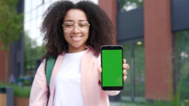 Üniversite binasının yanında duran yeşil ekranlı telefonu gösteren Afro-Amerikan kız öğrenci. Renk anahtarı, dikey model. Reklam şablonu. Genç kadın kameraya bakarak akıllı telefon gösteriyor.