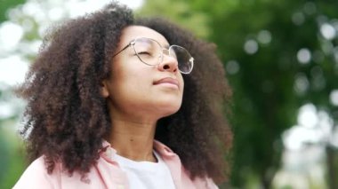 Şehir parkında gözleri kapalı duran mutlu Afro-Amerikan kadın. Gülümseyen siyah bir kadın öğrenci derin nefes alır ve doğada olup kameraya bakmaktan zevk alır. Kapat.