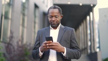 Olgun Afro-Amerikan iş adamı akıllı telefon kullanarak ofis binasının önünde dikiliyor. Resmi giyinmiş, gülümseyen, teknolojiyle uğraşan bir profesyonel. Modern şehir merkezi.