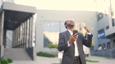Olgun Afro-Amerikan iş adamı ofis binasının önünde durmuş cep telefonundan iyi haberler okuyor. Takım elbiseli siyah adam başarıyı kutluyor. İş başarısını profesyonel başarıyı kabul et.