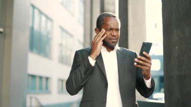 Takım elbiseli Afrikalı Amerikalı işadamı dışarıda kötü haberler alıyor. Adam akıllı telefonunu tutarken hayal kırıklığına uğramış görünüyor. Şehir ortamında vurgulanmış ifadeye sahip işadamı mesajı okuyor