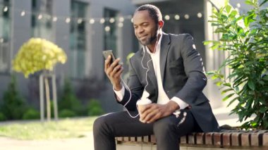 Takım elbiseli Afro-Amerikan bir adam dışarıda oturuyor, akıllı telefon kullanıyor ve kahve içiyor. Rahatlamış iş adamı dinleniyor, gülümsüyor ve kulaklıkla dinliyor. Ofis yakınlarındaki şehir merkezinde güneşli bir gün