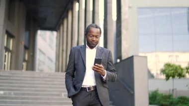 Afro-Amerikalı işadamı akıllı telefon kullanarak ofis binasının önünde yürüyor. Takım elbiseli profesyonel bir adam şirket binasının yakınındaki bir sokakta telefon teknolojisi kullanıyor. İş iletişimini kavra
