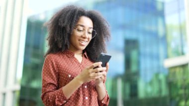 Gülümseyen Afro-Amerikan kadın ofis binasının dışında elinde akıllı telefonla başarıyı kutluyor. Mutlu zenci kadın profesyonel telefonda iyi haberler okuyor, heyecan ve neşe gösteriyor. Kapat.