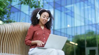 Modern ofis binasının dışında bankta oturan, kulaklık takan ve bilgisayarında video görüşmesi yapan Afro-Amerikalı bir kadın. Kendinden emin gülümseyen siyah kadın sanal bir sohbete daldı.