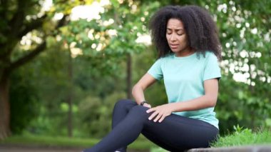 Genç Afro-Amerikan sporcu kadın şehir parkında kas ağrısıyla oturuyor. Siyah bir kadın masaj yapıyor, esneme hareketleri, travma yaralanmaları. Fitness dişisinin burkulması şiddetli ağrı germe kuvveti. Bacağa kramp giren baldır sporu