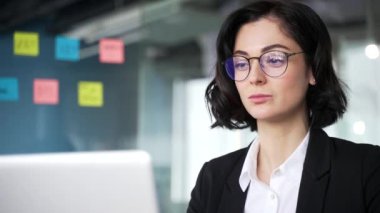 Çağdaş bir ofiste dizüstü bilgisayarla çalışan kendine güvenen bir kadın. İş kadını gözlük takıyor, görevlere ve projelere konsantre oluyor. Yenilikçi fikirleri olan modern çalışma alanı. Kapat.