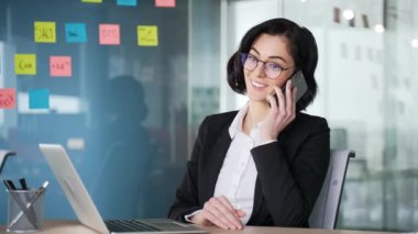 Ofisteki bir iş yerinde otururken telefonla konuşan kendine güvenen bir iş kadını. Bayan bir yönetici müşterisiyle konuşuyor. İş elbisesi içindeki profesyonel kadın birden fazla iş yapıyor, iletişim kuruyor.