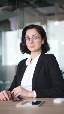 Kendine güvenen gözlüklü bir iş kadını modern bir ofiste çalışma masasında oturuyor. İş kıyafeti giymiş, kısa siyah saçlı profesyonel bir kadın. Kadın yöneticinin vesikalığı. Dikey video