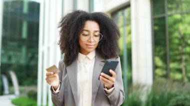 Modern ofis binasının dışında kredi kartı ve akıllı telefon tutan Afrikalı bir iş kadını. Mutlu gülümseyen siyah kadın online alışveriş yapıyor telefon, e-ticaret, mutlu ve başarılı bir alıcı kullanıyor.
