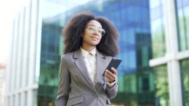 Modern ofis binasının dışında akıllı telefon kullanan kendine güvenen Afrikalı Amerikalı iş kadını. Takım elbiseli profesyonel bir kadın iş sırasında mesajları veya güncellemeleri kontrol ediyor. İş anlayışı, teknoloji
